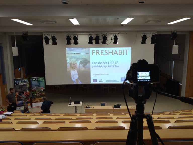 Freshabit LIFE IP yhteistyötä ja toimintaa dianäkymä kuvattuna Ilomantsin Kunnantalon Kalevala-salissa yläriville sijoitetun videokameran takaa.